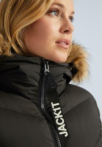 Femeie blondă îmbrăcată într-o jachetă de iarnă neagră cu umplutură, cu glugă căptușită cu blană, prim-plan al fermoarului jachetei etichetat "JACK1T."