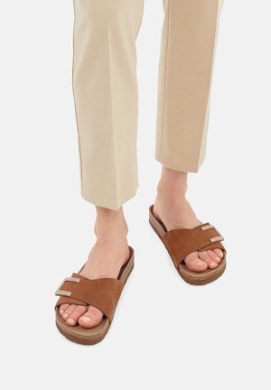 Sandalete din suede maro, cu două accesorii metalice cu cataramă și o talpă texturată din plută, asortate cu pantaloni bej, ajustați.