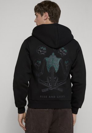 Junger Mann trägt schwarzen Hoodie mit blau akzentuiertem Skelett- und Totenkopf-Design und dem Text „RISE AND GRIND“ auf dem Rücken, seitlich stehend.