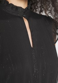 Blouse noire avec un col à volants, présentant une découpe en forme de goutte d'eau. Le tissu a une surface texturée avec des détails subtilement scintillants.