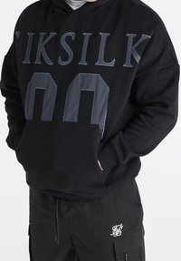 Felpa nera con grande scritta "IKSIL" e design doppio "00" sulla parte anteriore. Presenta una tasca a marsupio e polsini a costine. Indossata con pantaloni cargo neri.