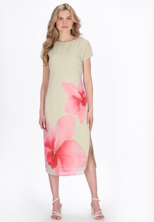 Blondynka w beżowej sukience midi z dużymi różowymi kwiatowymi wzorami i rozcięciem po bokach, stoi na białym tle w białych sandałach na obcasie.