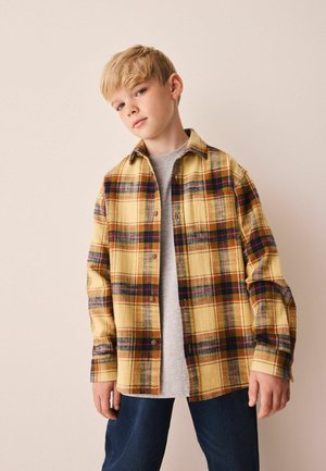 Blond chłopiec w żółtej koszuli w kratę na szarym t-shircie i niebieskich dżinsach, stojący na jednolitym jasnym tle.