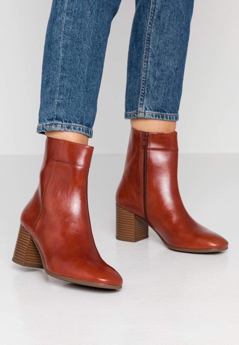 Femme portant un jean bleu moyen et des bottines en cuir marron à talons blocs avec zips latéraux, debout sur un sol blanc lisse.