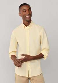 Hombre sonriente con camisa amarilla clara de manga larga remangada y pantalones beige, de pie frente a un fondo liso de color gris claro.