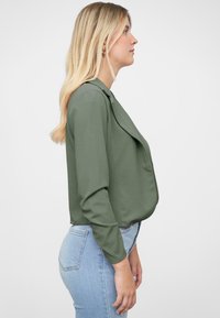 Blusa de color verde oliva con un frente drapeado, mangas largas y una textura suave, combinada con jeans azules claros. Diseño sencillo y forma ajustada.