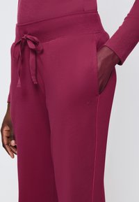 Bordeaux joggers met een zachte textuur, voorzien van een strikceintuur, zijzakken en contrasterende roze stiksels. Kleine logo-detail op de heup.