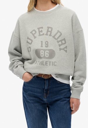 Kobieta w jasnoszarej bluzie Superdry z napisem "1986 Athletic", w połączeniu z niebieskimi dżinsami i plecionym brązowym paskiem, ręka w kieszeni.