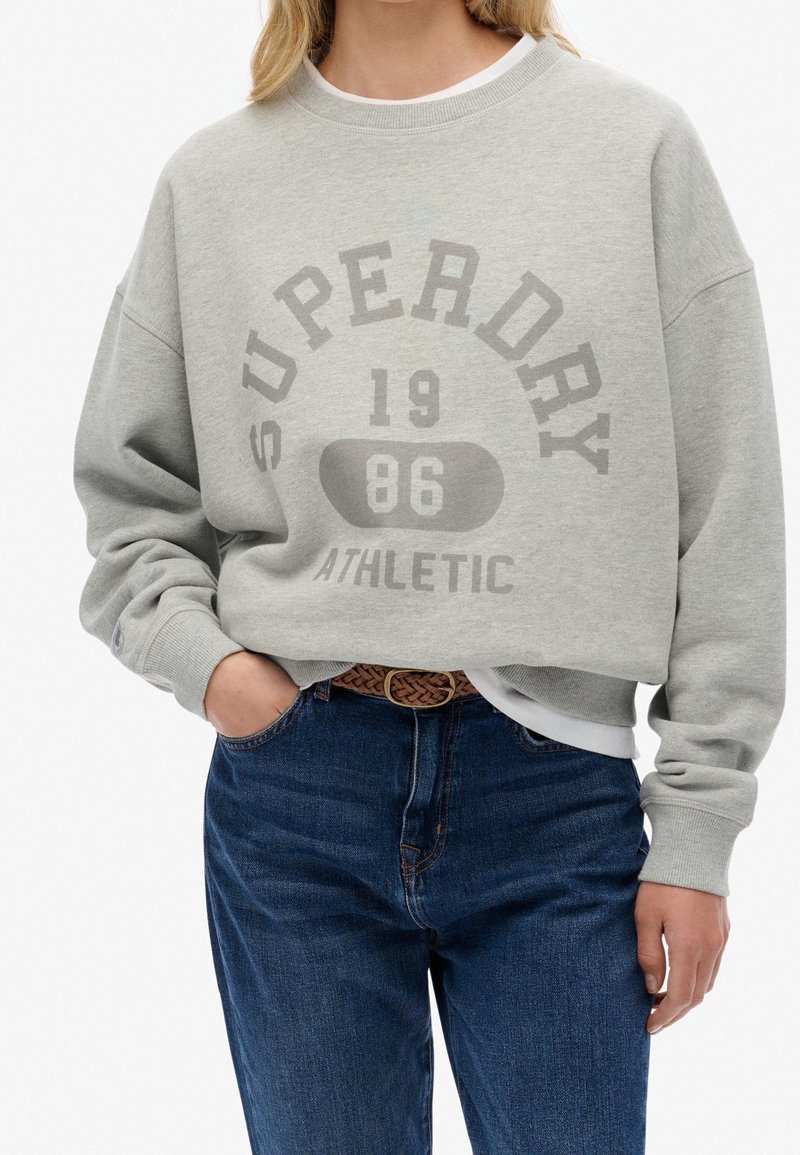 Femme portant un sweat-shirt gris clair Superdry avec le texte "1986 Athletic", associé à un jean bleu et une ceinture marron tissée, main dans la poche.