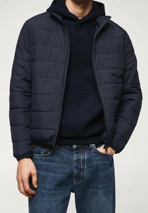 Homme portant une veste matelassée bleu marine par-dessus un sweat à capuche noir avec un jean bleu, debout avec une main dans la poche devant un fond uni.