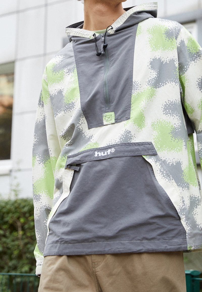 Homme portant une veste à capuche avec un motif camouflage vert, gris et blanc et une poche avant étiquetée "huf" à l'extérieur près d’un bâtiment.