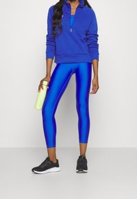 Sudadera atlética azul con media cremallera, combinada con leggings azules brillantes. Zapatillas deportivas negras. Persona sosteniendo una botella de agua verde claro.
