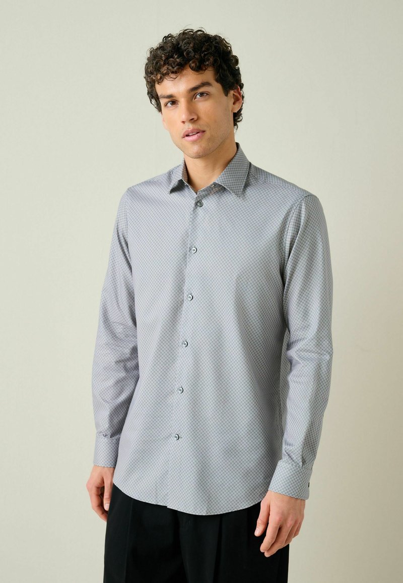 Jeune homme aux cheveux bouclés portant une chemise à manches longues boutonnée gris clair à motifs et un pantalon noir sur un fond neutre.
