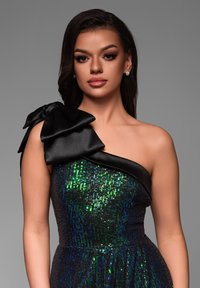 Svart enaxlad klänning med en stor satängsrosett och glittrande gröna och blå paljetter på bysten, som visar en texturerad tyg.
