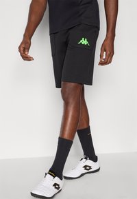 Fekete sportos rövidnadrág zöld Kappa logóval, oldalzsebekkel és laza szabással. Fehér cipőkkel és fekete mintás zoknival párosítva.