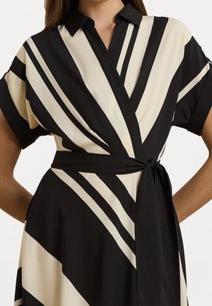 Femme portant une robe portefeuille rayée noire et beige avec manches courtes, col et nœud à la taille noir.