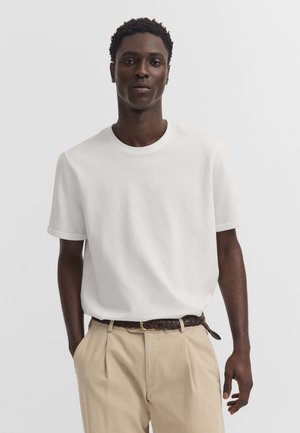 Mann trägt ein schlichtes weißes kurzärmeliges T-Shirt, das in beige Hose mit braunem geflochtenem Gürtel gesteckt ist, steht mit einer Hand in der Tasche.