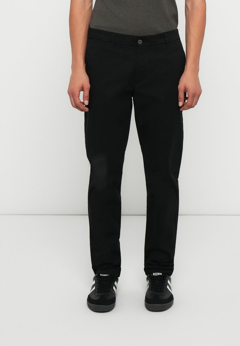 Pantaloni in cotone neri con taglio slim, dotati di chiusura frontale con bottone, tasche laterali e una texture liscia. Indossati con sneakers nere.