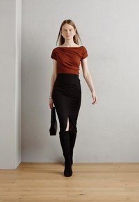 Brun drapert topp, svart tettsittende skjørt med sidesliss, og knelhoye svarte støvler. Modellen holder en svart veske, står på tregulv.