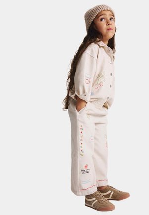 Chemise et pantalon beige brodés avec des motifs floraux et fruitiers. Portés avec un bonnet tricoté marron clair et des baskets marron.