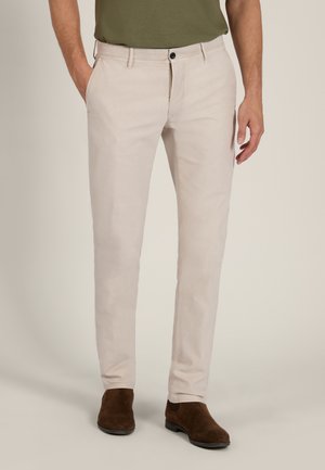 Mann trägt beige Slim-Fit-Hose, olivgrünes Hemd und braune Wildleder-Stiefeletten, steht mit einer Hand in der Tasche.