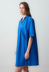 Abito camicia blu con maniche corte arrotolate, colletto e dettaglio a pieghe sulla parte anteriore. Realizzato in tessuto liscio per una vestibilità comoda.