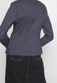 Blazer marine avec des rayures verticales blanches, au design ajusté et en tissu lisse. Présente une silhouette taillée et un ourlet légèrement courbé au niveau de la taille.
