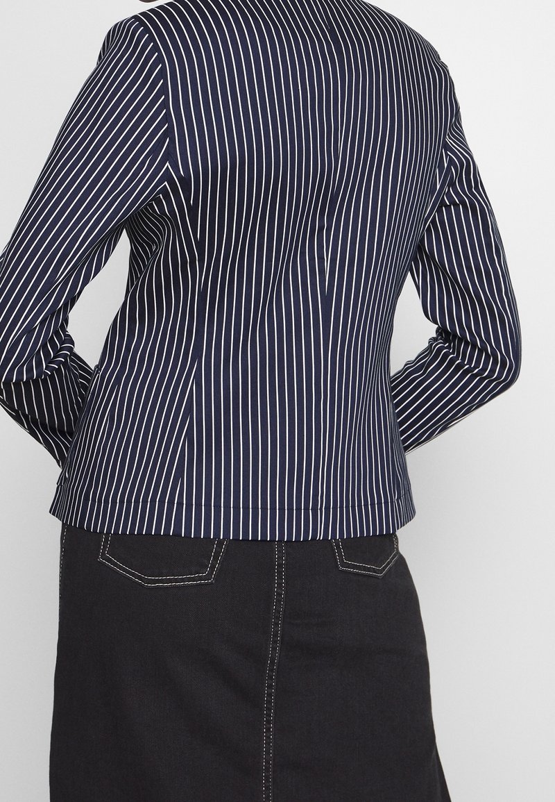 Blazer marine avec des rayures verticales blanches, au design ajusté et en tissu lisse. Présente une silhouette taillée et un ourlet légèrement courbé au niveau de la taille.