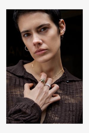 Anel dourado em forma de coração com texto, anel prateado estriado, colar de ouro em camadas e camisa castanha texturizada. Close-up da mão e do rosto.