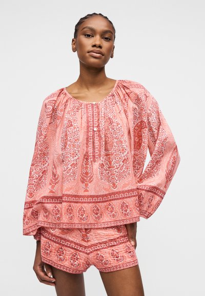 Femme portant un chemisier ample à manches longues à motifs roses et un short assorti, debout avec une main détendue le long du corps.
