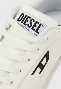 Fehér bőr sportcipő szürke fűzőkkel, fekete hímzett „A” logóval és egy „DIESEL” feliratot tartalmazó fekete címkével fehér alapon.