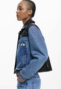 Young person with braided hair wears blue denim jacket with black leather shoulders and matching jeans with cartoon patches.