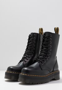 Botas de cuero negro con cordones, con una plataforma gruesa y suela texturizada, que cuentan con una lengüeta amarilla con marca y ojales de metal.