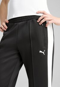 Czarne sportowe spodnie z fakturowanej tkaniny, białe boczne paski, elastyczny pasek w talii oraz kieszeń na zamek. Posiadają małe białe logo Pumy.
