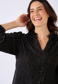 Blusa nera con texture e strisce dorate verticali, dotata di colletto alto e chiusura con bottoni, con maniche a sbuffo.