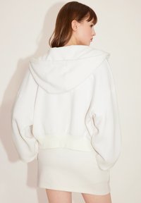 Sudadera con capucha blanca de corte relajado, que cuenta con una capucha grande, mangas anchas y un cinturón de canalé. Textura suave y diseño minimalista.