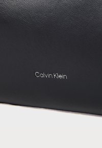 Clutch in pelle nera con finitura testurizzata, dotata di un logo "Calvin Klein" in argento nell'angolo in basso a destra.