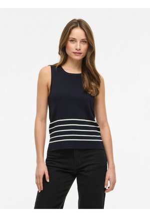 Ženska z dolgimi rjavimi lasmi, oblečena v brezrokavnik temno modre barve z belimi vodoravnimi črtami blizu roba in v črnih hlačah, stoji pred belim ozadjem.