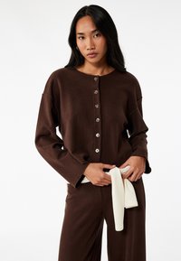 Brun knäppskjorta med långa ärmar och vida manschetter, prydd med stora knappar. Matchas med matchande byxor, en krämfärgad slips är knuten runt midjan.