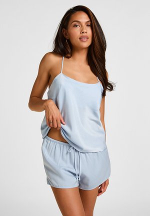 Femme portant une camisole en satin bleu clair et un short assorti, debout devant un fond gris uni, tenant l'ourlet de la camisole d'une main.