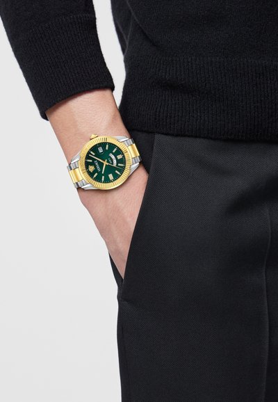 Kultainen ja hopeinen rannekello, jossa on vihreä taulu, päivämäärätoiminto ja rohkeat tuntimerkinnät. Käytetty ranteessa, yhdistetty mustien housujen kanssa.