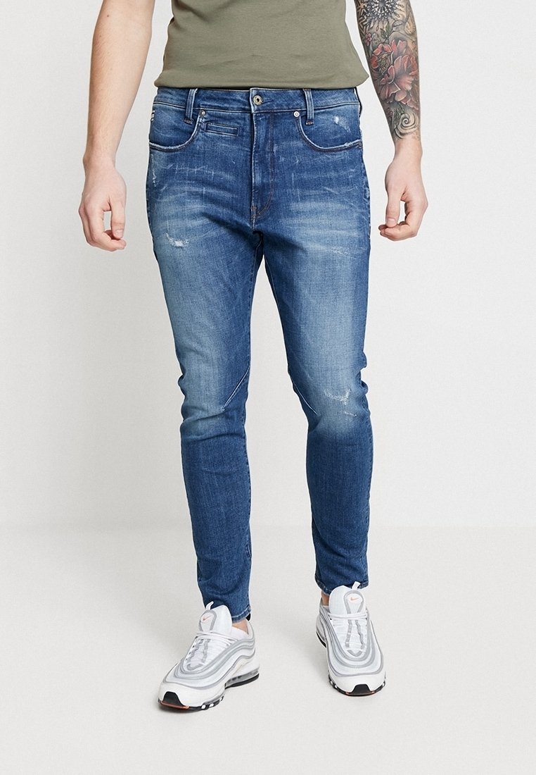 Homme portant un jean slim bleu délavé, des baskets blanches et une chemise vert olive, debout devant un fond blanc uni.