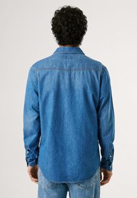 Blå denimskjorta med långa ärmar, knappmanschetter och spetsig krage. Har en något rundad fåll och en sydd bakåkt.