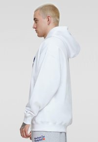 Witte pullover hoodie met een ruime pasvorm en een grote capuchon, kenmerken zijn ribgebreide manchetten en een relaxed fit. Gdrag met lichtgrijze sweatpants.