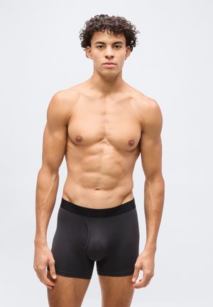 Junger Mann mit lockigem Haar steht frontseitig, oberkörperfrei, trägt schwarze Boxershorts vor schlichtem weißem Hintergrund.