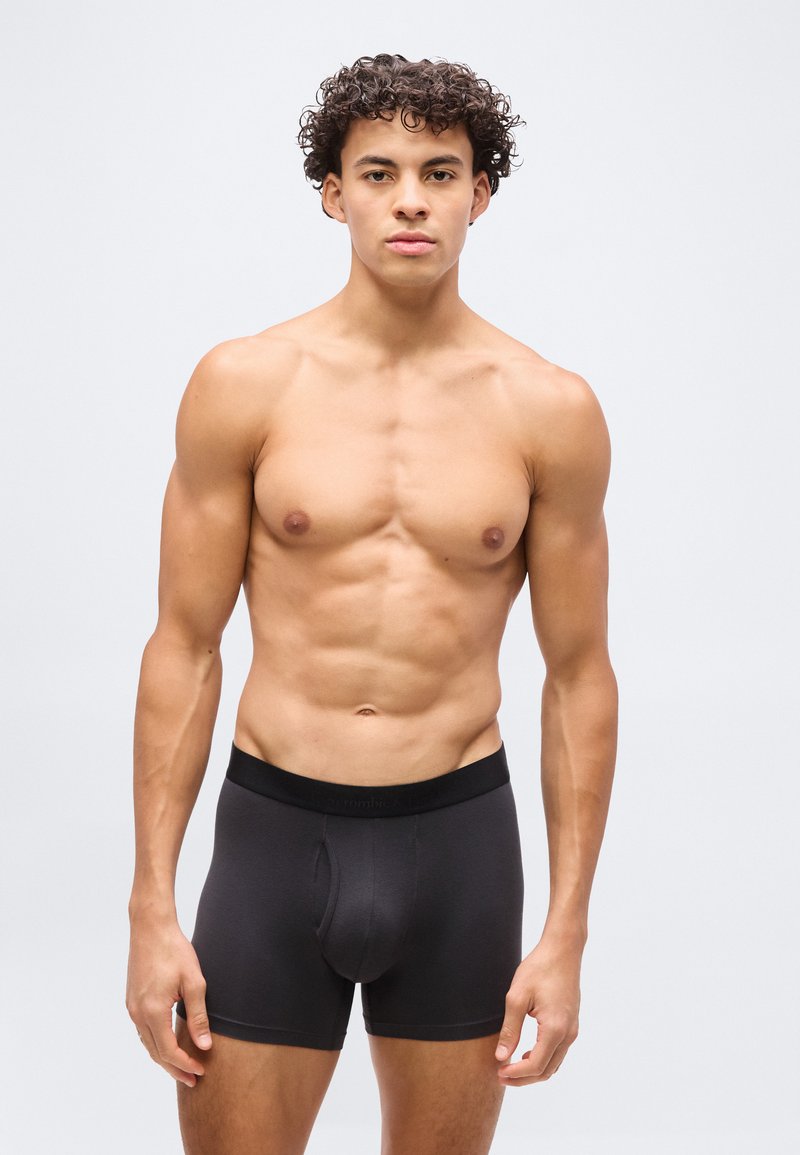 Νεαρός άνδρας με σγουρά μαλλιά στέκεται αντίκρυ, χωρίς πουκάμισο, φορώντας μαύρα boxer briefs σε απλό λευκό φόντο.