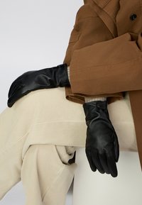 Sorte læderhandsker med en glat tekstur, der har et tætsiddende design og detaljer ved håndleddet, båret over et beige stofoutfit.