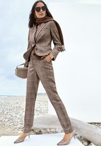 Braunes kariertes Anzug mit einem taillierten Jacket und schmal zulaufenden Hosen, kombiniert mit einem braunen Schal und Schuhen im Schlangenleder-Muster. Die Accessoires umfassen eine Sonnenbrille.