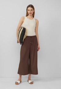 Débardeur côtelé beige clair associé à un pantalon large marron, doté d'une taille élastique, et des sandales blanches décontractées avec des accents dorés.