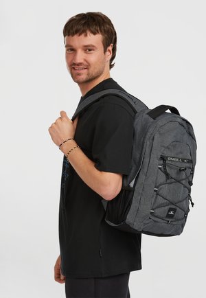 Mochila gris con acentos negros, tela texturizada, correas ajustables, detalles de cuerda elástica y bolsillo de malla lateral, llevada por un hombre.